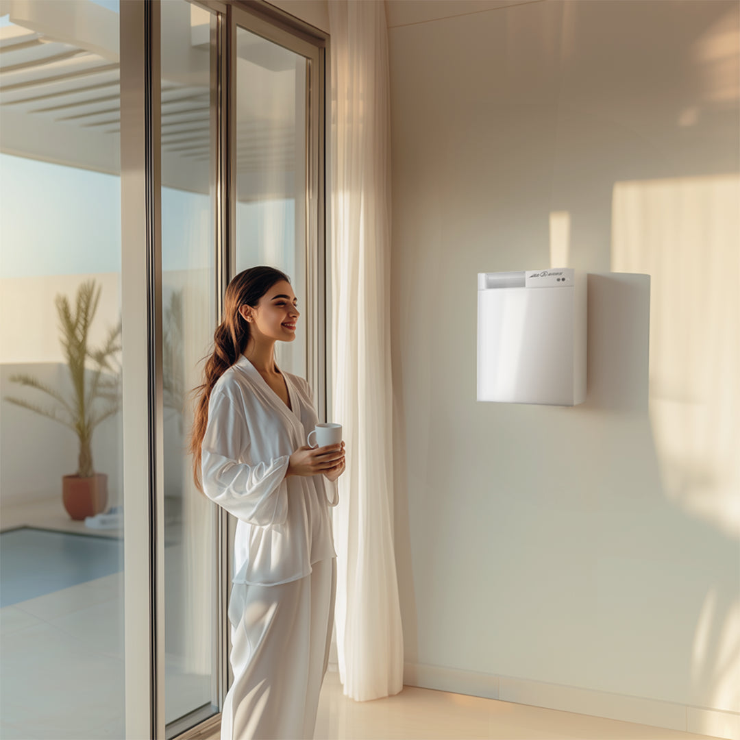 Woman in a white robe standing by a window holding a cup, with a white air purifier on the wall.